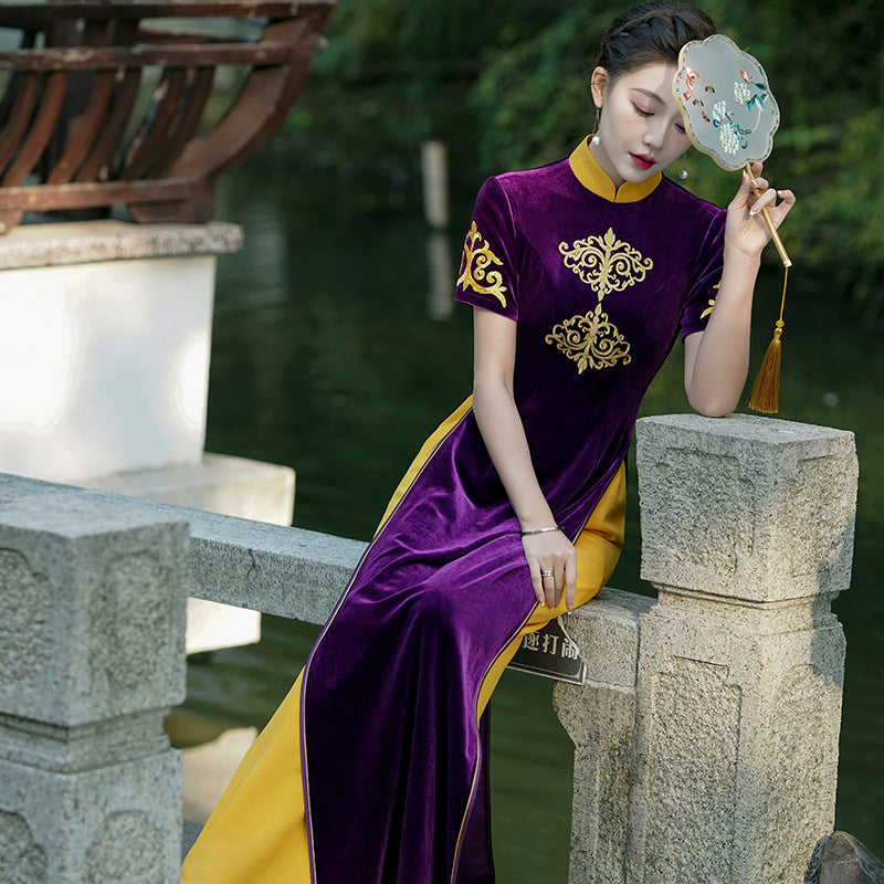 Full-body seated portrait of a model wearing the Imperial Plum Traditional Ao Dai Dress in deep purple velvet with gold embroidered motifs. The Vietnamese ao dai dress features a high collar, fitted silhouette, and contrasting golden side panels. Photographed outdoors beside a stone bridge and water, creating an elegant and regal modern ao dai aesthetic.