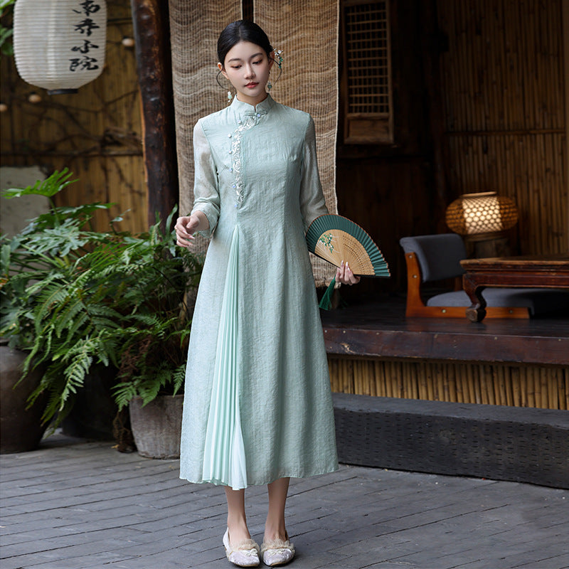 Frontal full-body view of a woman wearing a mint green modern Ao Dai dress with long sleeves and embroidered collar, standing in soft outdoor lighting near a wooden tea house.