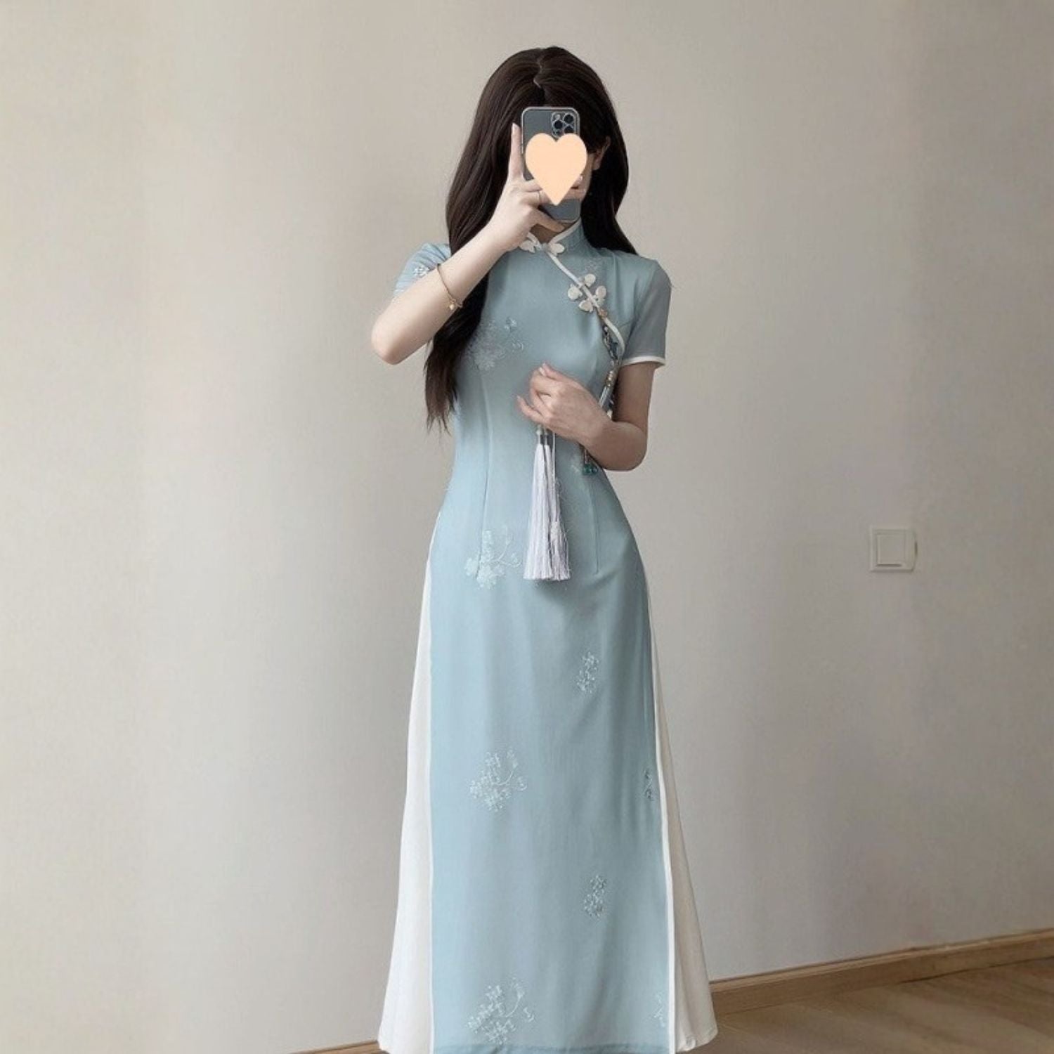Front-angle full-body shot of a woman wearing a modern Vietnamese Ao Dai dress in pastel blue with subtle floral embroidery and white side panels, and holding a tassel. Captured indoors under soft neutral lighting with minimal background.