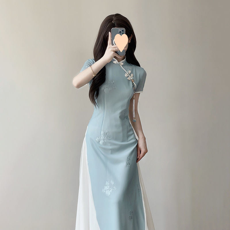 Front-view body shot of a woman wearing a modern Vietnamese Ao Dai dress in light blue with embroidered floral patterns and white side panels, photographed indoors with soft neutral lighting.