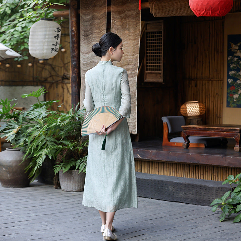 Back view of a woman in a light green Ao Dai dress holding a folded fan behind her, standing in front of a traditional wooden tea house in natural daylight.