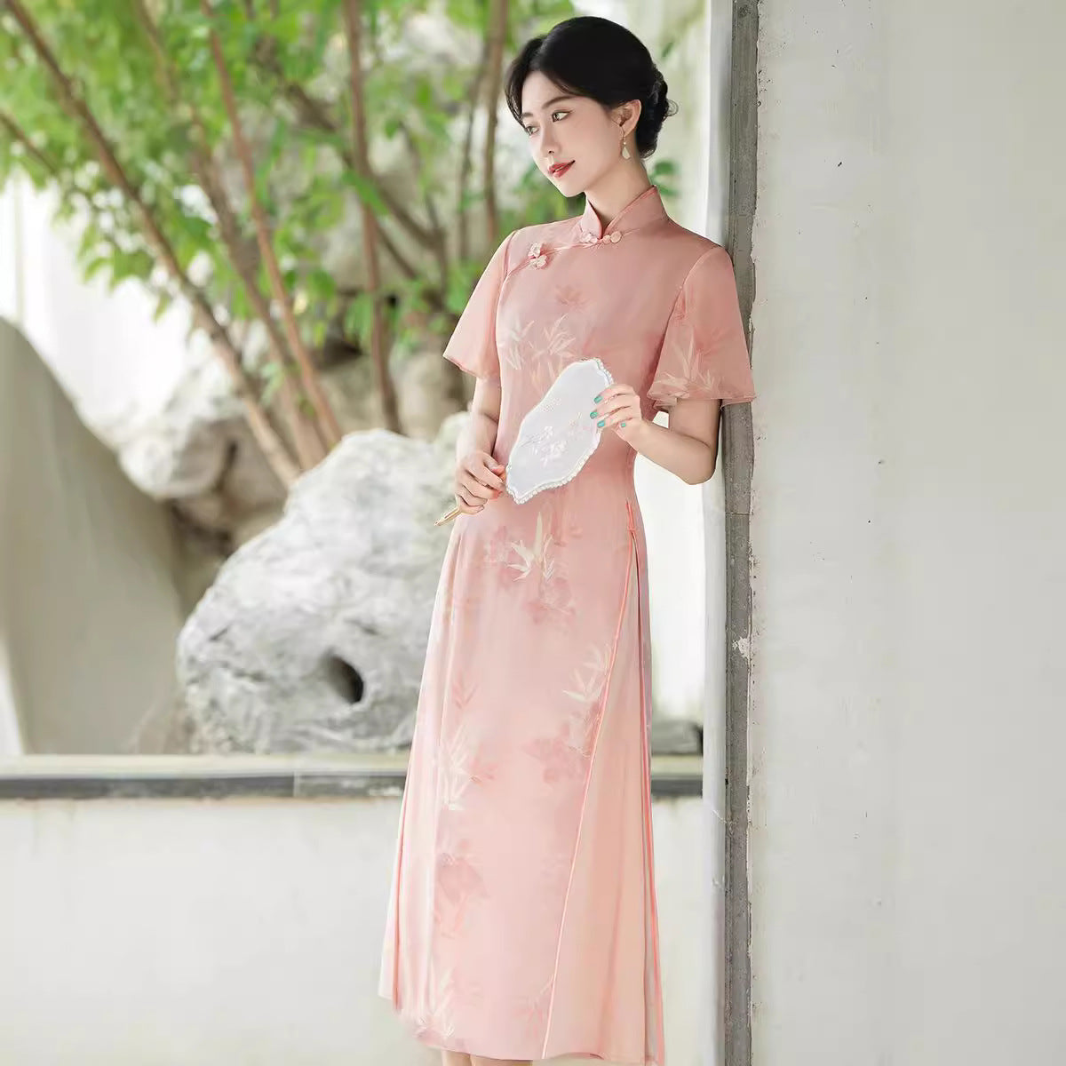 Three-quarter leaning pose of a woman holding a white lace fan while modeling the Pink Serenity Traditional Ao Dai. The Vietnamese Ao Dai’s bamboo print and blush tone create a serene, timeless look.