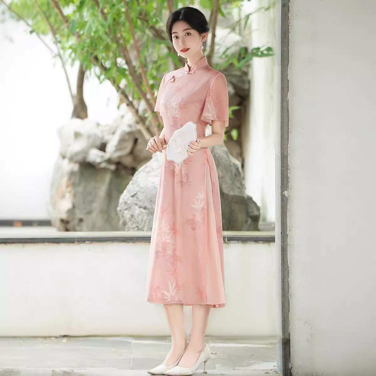 Side-angle portrait of a woman in the Pink Serenity Traditional Ao Dai, resting lightly against a wall. The Ao Dai’s pink silk fabric and flutter sleeves shine under natural daylight.