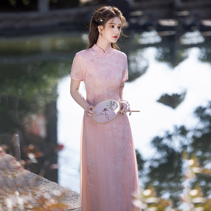 Outdoor close-to-full-body shot of the Silken Lake Modern Ao Dai Dress in pink, woman holding a round embroidered fan beside a calm water surface; subtle bamboo-inspired pattern; refined, modern Vietnamese ao dai styling.