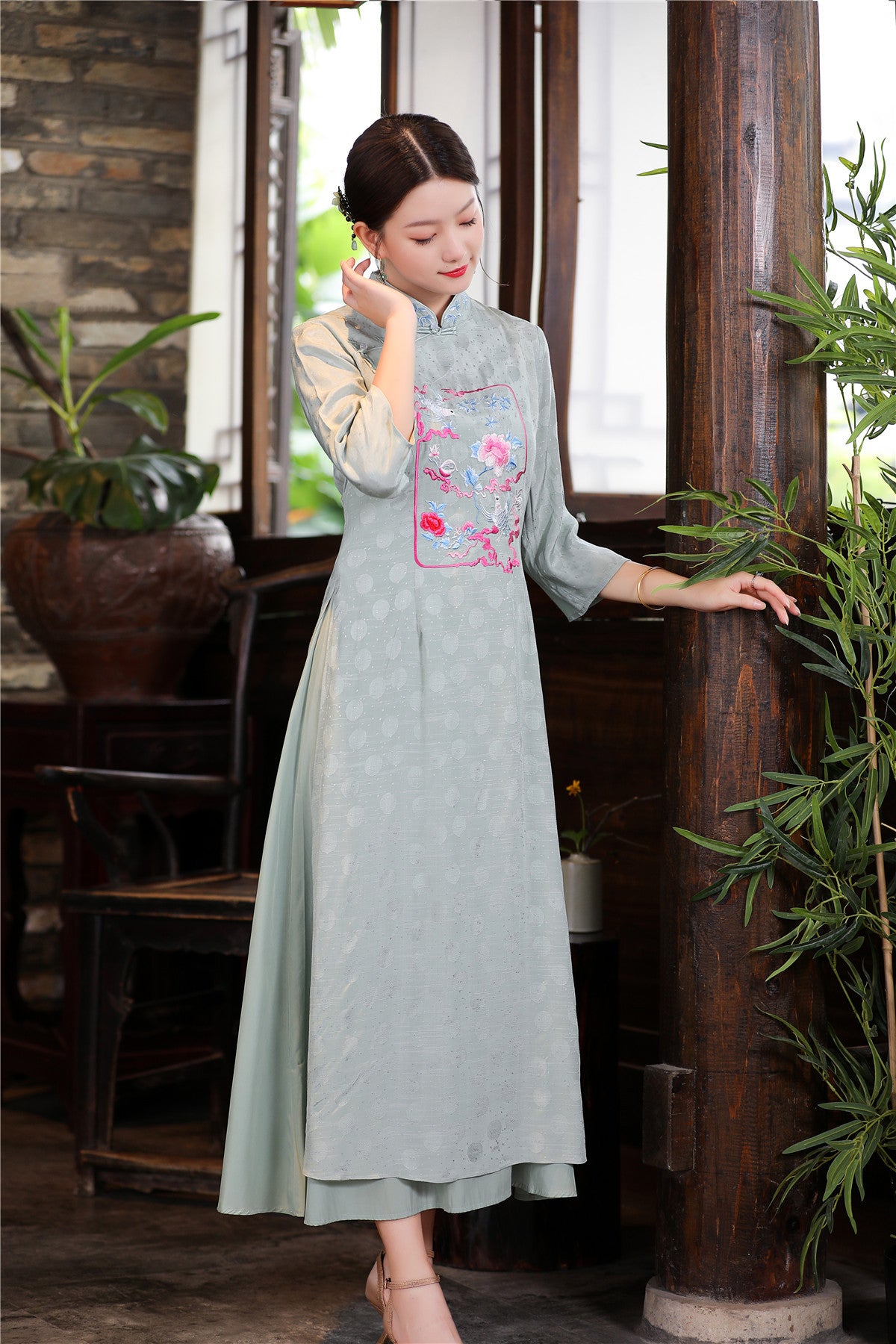 Model wearing sage green traditional Ao Dai with embroidered floral chest panel, standing indoors by wooden window, showcasing elegant cultural craftsmanship.
