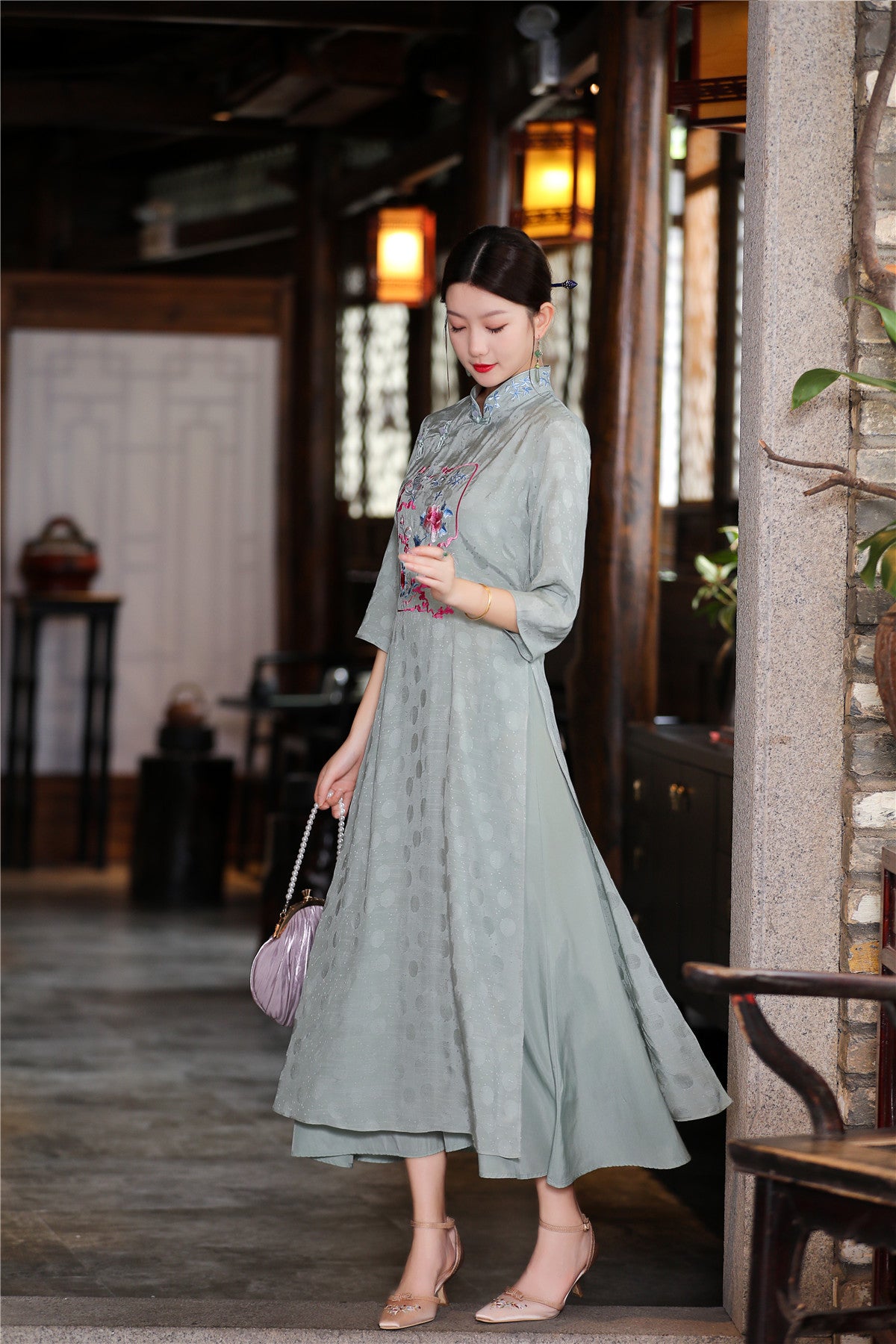 ull-length view of sage green traditional Ao Dai dress with mid-length sleeves and layered skirt, model holding a handbag in a classical Vietnamese interior setting.

SEO Filename: