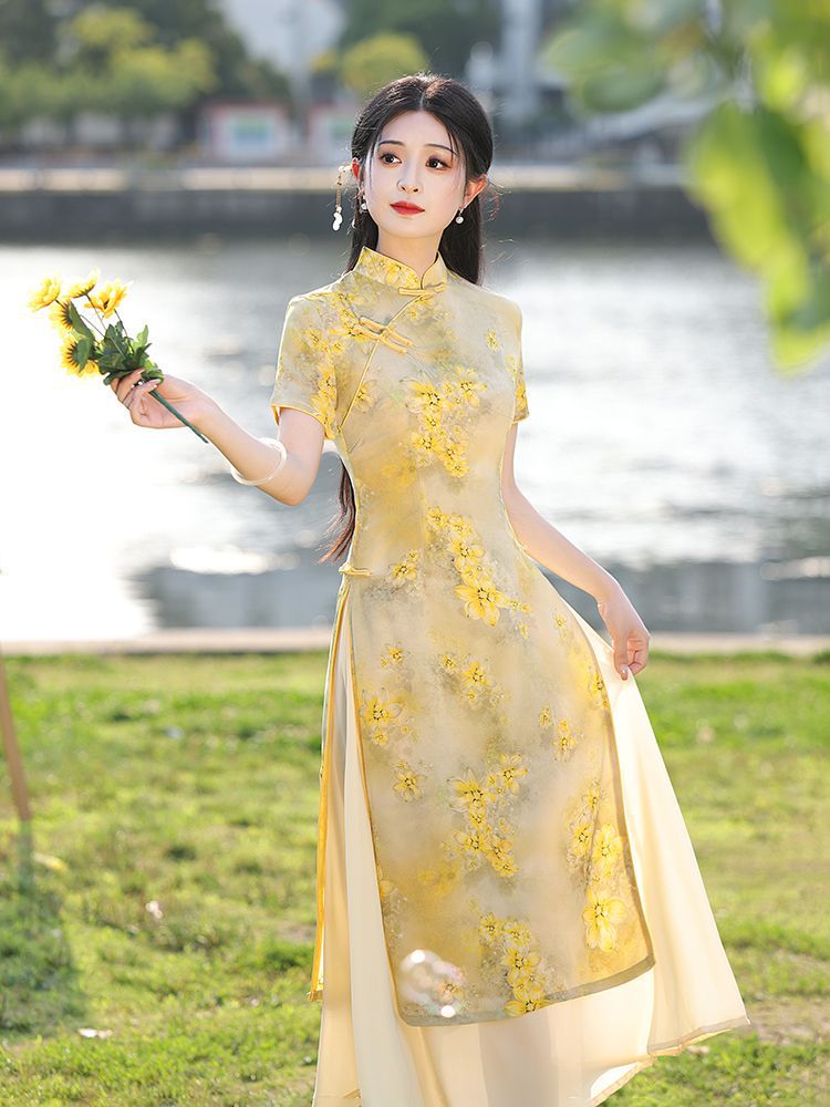 Elegant standing pose of a woman wearing a light golden Vietnamese ao dai dress with layered chiffon panels, photographed in a park by the water.