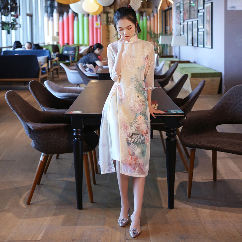 Full-length portrait of a woman standing between tables in a café, modeling the Victoria Grade Modern Ao Dai Dress. The cream floral design and lace cuffs reflect refined Vietnamese modern fashion.