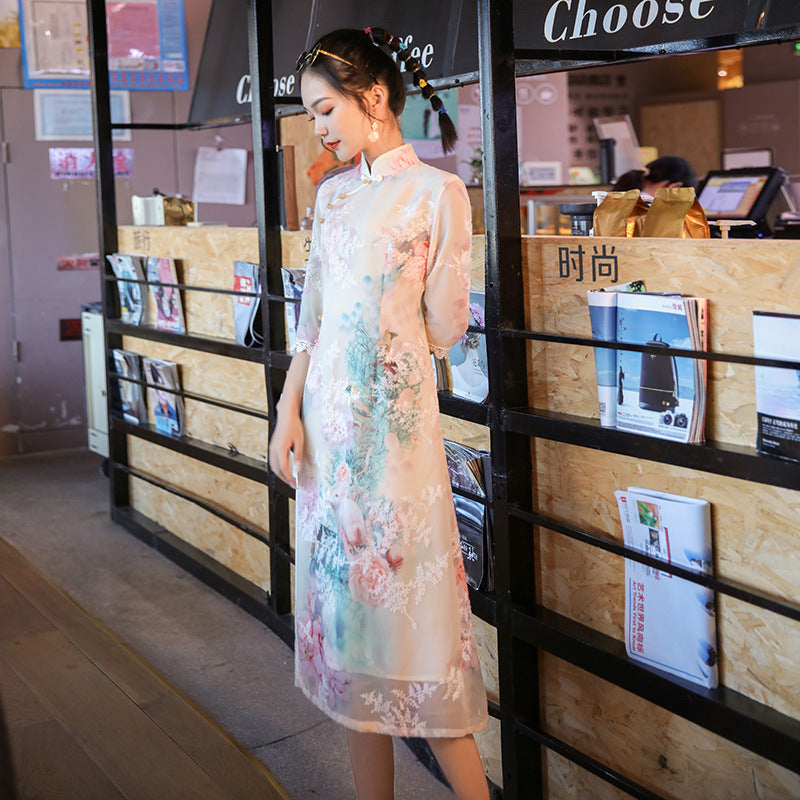 Three-quarter angle shot of a woman wearing the Victoria Grade Modern Ao Dai Dress in soft pastel chiffon, photographed in a cozy café interior. The modern Ao Dai blends Vietnamese heritage with a graceful feminine silhouette.